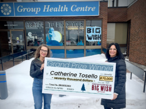 Two women hold a giant presentation cheque in front of Group Health Centre Building in the winter