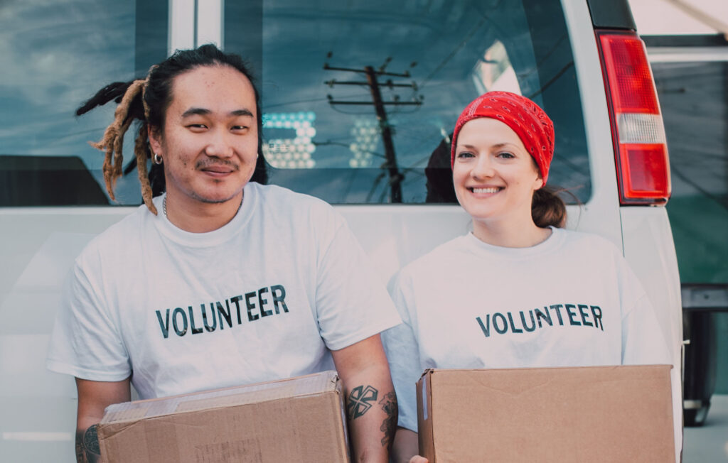 two people stand holding cardboard boxes while wearing a white shirt that says Volunteer on it. The right person is wearing a red bandana